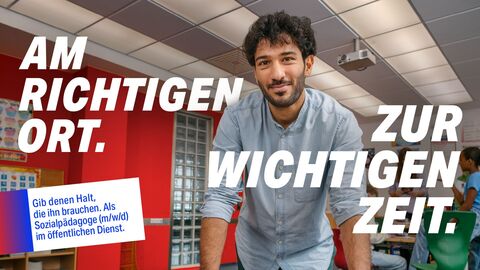 Gebe denen Halt, die ihn brauchen. Bewirb dich für einen sozialen Beruf. Motiv Sozialpädagoge: Ein junger Mann mit Bart steht in einem Klassenzimmer. Im Hintergrund sind Tische, Stühle und eine Tafel zu sehen. Der Text auf dem Bild lautet: "AM RICHTIGEN ORT. ZUR WICHTIGEN ZEIT." und "Gib denen Halt, die ihn brauchen. Als Sozialpädagoge (m/w/d) im öffentlichen Dienst."